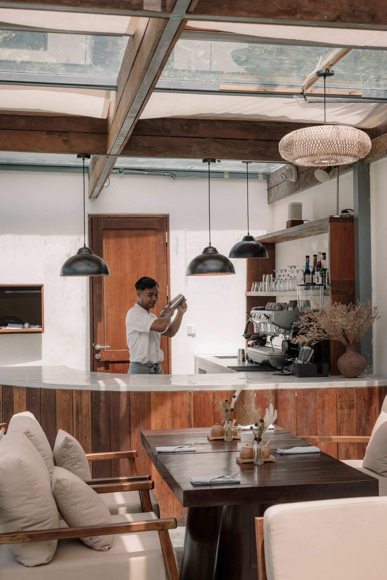 The barista shaking a drink behind Bonnie’s curved teak bar, linen armchairs and a laid table in front.