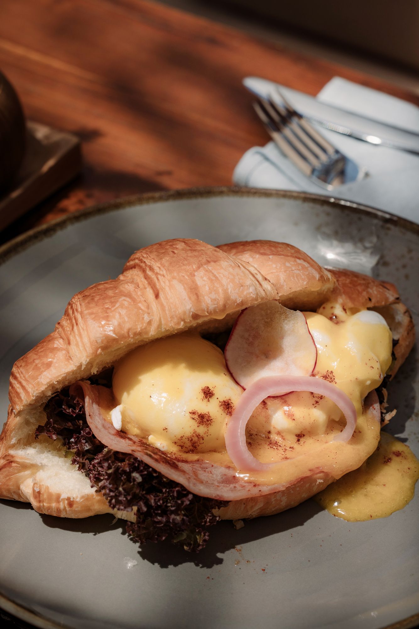 A croissant split and filled with ham, poached eggs and hollandaise, red onion on top.