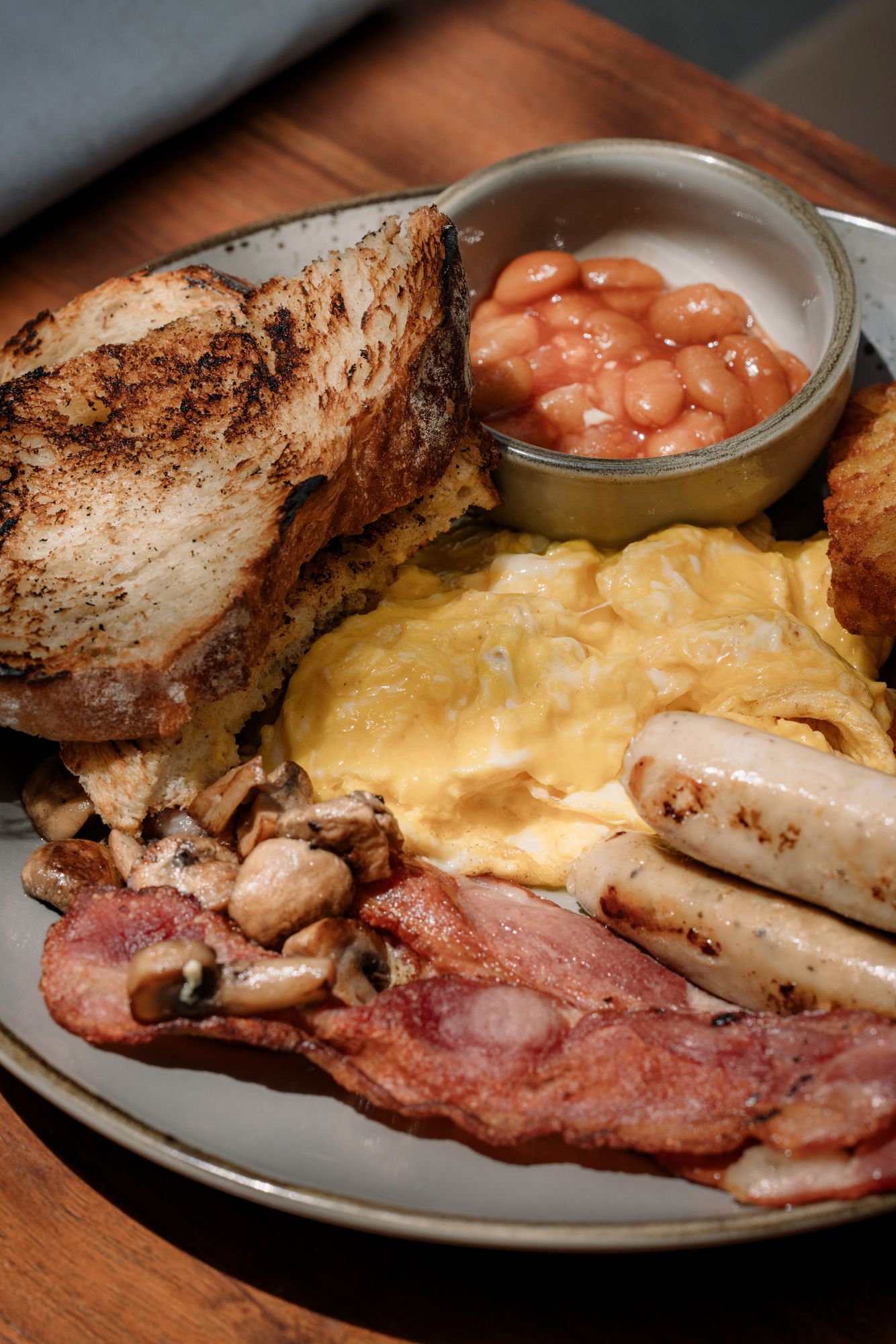 A full breakfast of scrambled egg, sausages, bacon, mushrooms, hash browns, toasted sourdough and a pot of baked beans.