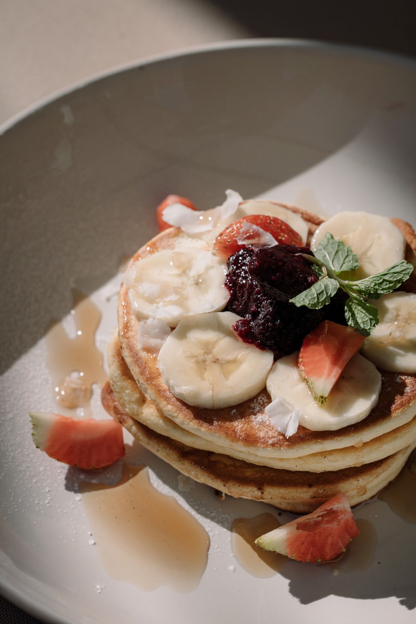 A stack of pancakes with banana, strawberries, berry compote and mint, maple syrup pooling on the plate.