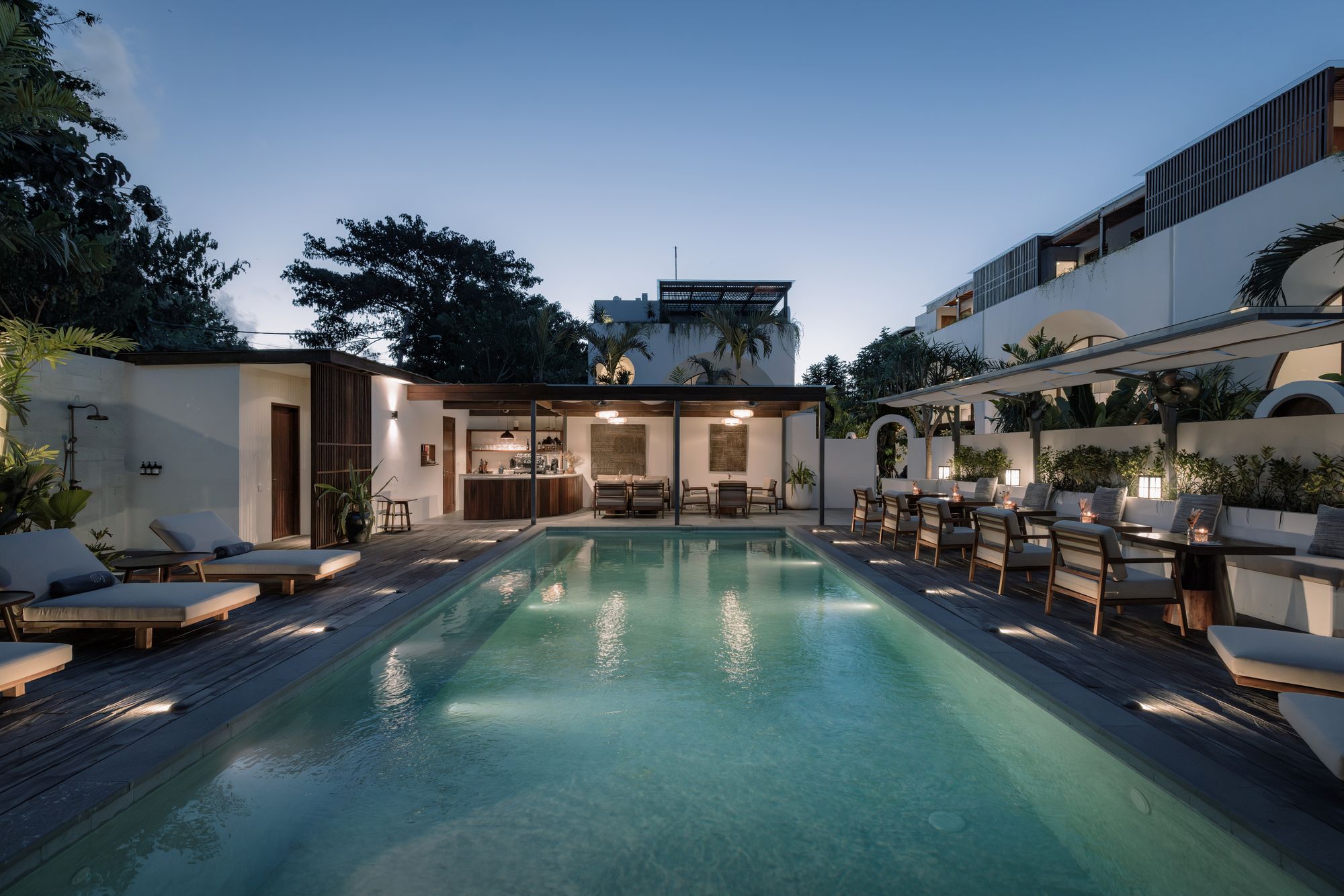 The pool lit from below at dusk, loungers and candlelit tables along the deck, the villas glowing above.