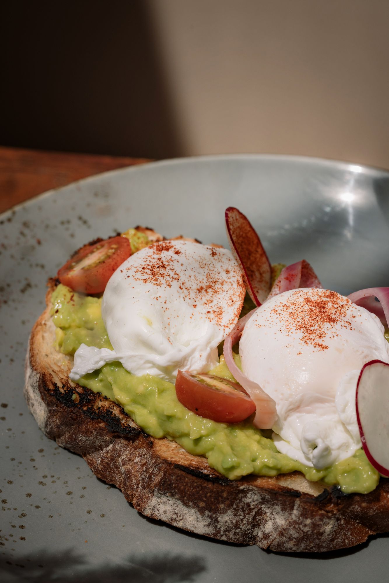 Smashed avocado on sourdough with two poached eggs, cherry tomatoes, radish and paprika on a speckled grey plate.