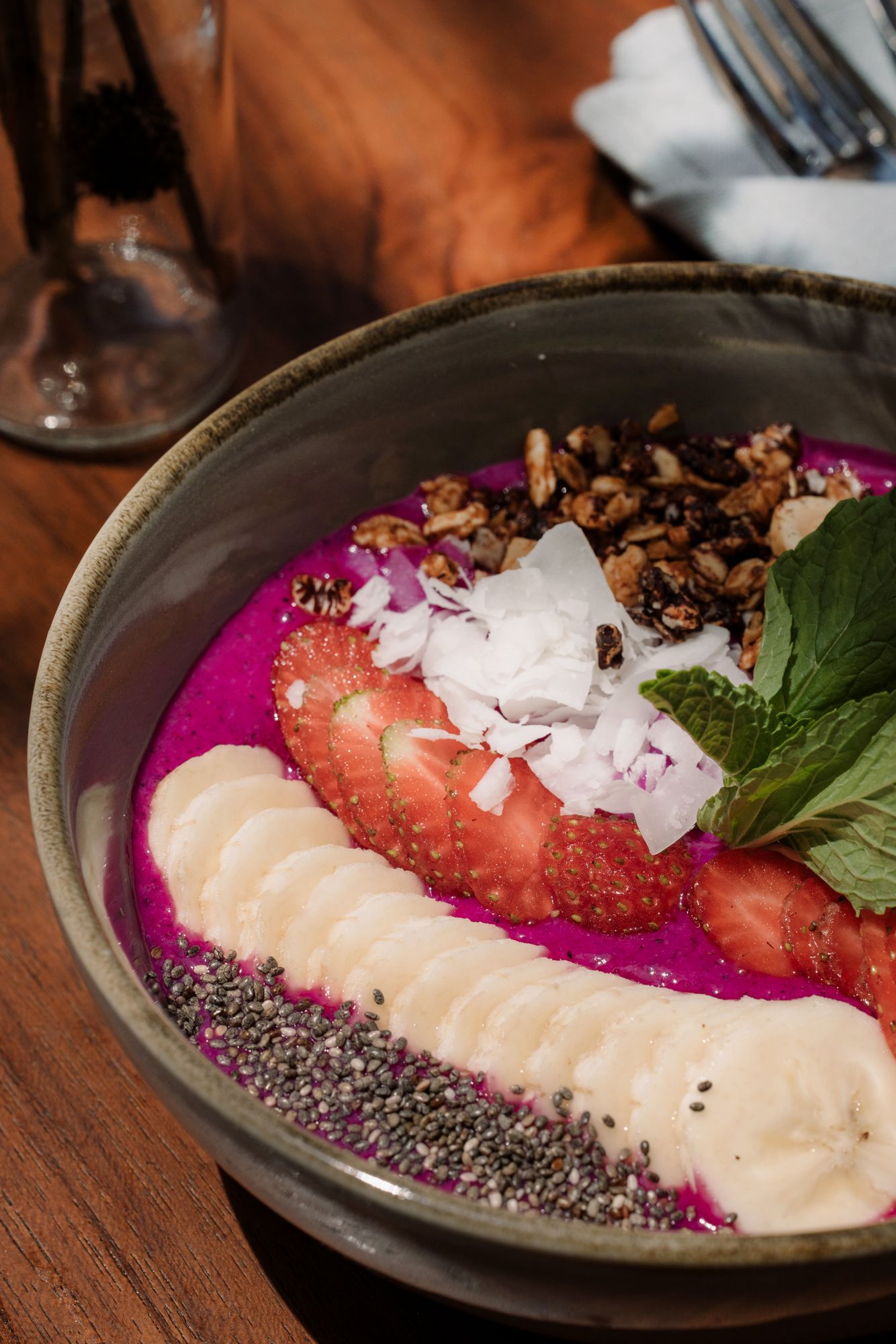 A bright pink dragon fruit smoothie bowl topped with banana, strawberries, granola, chia, coconut and mint.